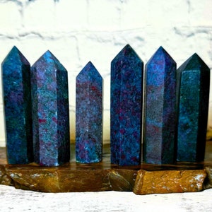 May include: Six polished gemstone towers with a mix of blue, purple, and red hues. The crystals are displayed on a brown wooden base against a white brick wall. The towers vary in size and shape.