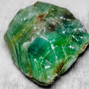 May include: A raw, uncut emerald gemstone with a vibrant green hue. The surface of the stone has a crystalline structure with areas of lighter and darker green, and some brown inclusions. The gemstone is irregularly shaped.