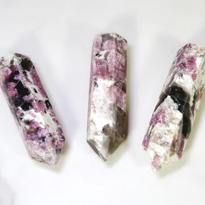 Pu&ograve; includere: Tre bacchette di cristallo di tormalina lucidata. I cristalli sono un mix di rosa, nero e bianco, con un'estremit&agrave; sfaccettata e appuntita. Le bacchette sono esposte su una superficie bianca.