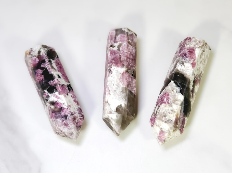 Pu&ograve; includere: Tre bacchette di cristallo di tormalina lucidata. I cristalli sono un mix di rosa, nero e bianco, con un'estremit&agrave; sfaccettata e appuntita. Le bacchette sono esposte su una superficie bianca.
