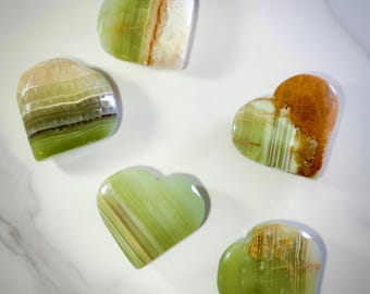 May include: Five heart-shaped stones in shades of green, brown, and white are arranged on a white surface. The stones have a polished, translucent appearance, with visible layers and patterns. The hearts vary in color and size.