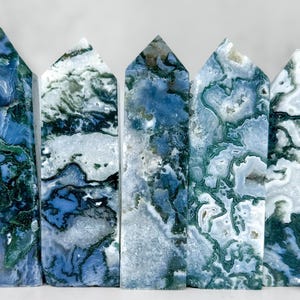May include: Five polished moss agate obelisks. Each obelisk is rectangular with a pointed top. The stones display a mix of blue, white, and green patterns. The obelisks are arranged in a row, showcasing their unique natural designs.