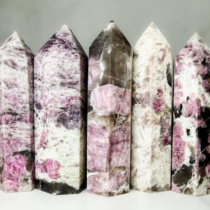 May include: Five polished crystal towers with pointed tops. The crystals display a mix of colors, including pink, white, and black. The crystals are arranged in a row, showcasing their unique patterns and textures. The crystals are approximately 5 inches tall.