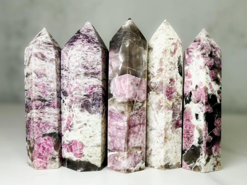 Pu&ograve; includere: Cinque torri di cristallo lucido con punte. I cristalli mostrano un mix di colori, tra cui rosa, bianco e nero. I cristalli sono disposti in fila, mostrando i loro motivi e le loro trame uniche. I cristalli sono alti circa 13 cm.