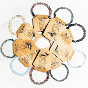 May include: A collection of beaded bracelets in various colors, including pink, green, and blue, arranged around brown tags. The tags feature illustrations and the text "VIA THE VEIL CRYSTAL KIDS".