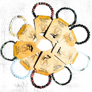 May include: A collection of beaded bracelets in various colors, including pink, green, black, and blue, arranged around several tan tags. The tags feature a celestial design and the text "VIA THE VEIL CRYSTAL KIDS."