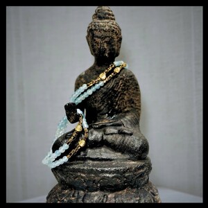 Puede incluir: Un collar de cuentas azules y doradas drapeado sobre una estatua de Buda de piedra gris oscuro. La estatua est&aacute; sentada en una postura meditativa con las manos en el regazo.