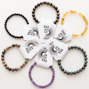May include: A collection of beaded bracelets in various colors, including black, brown, green, yellow, and purple. Each bracelet is displayed with a white tag that reads "VIA THE VEIL" in black text. The bracelets are arranged in a circular pattern.