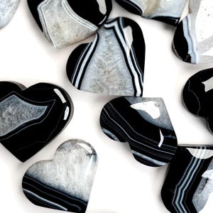May include: A collection of polished heart-shaped stones featuring banded patterns of black, white, and translucent grey. Each stone displays a unique combination of dark opaque layers and crystalline centres with a smooth, reflective surface finish.
