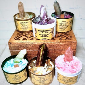 May include: Six candles in glass jars with gold lids, each with a different crystal and a label with a different affirmation. The affirmations are "Earth Mama", "Awaken Dreamer", "Manifest Rainbows & Joy", "I AM", "Roar Girl Roar", and "Sorbet Love".