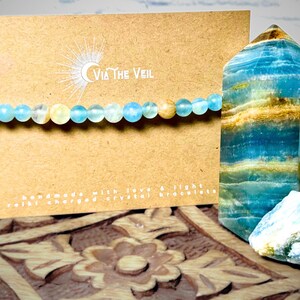 Puede incluir: Un brazalete de cristal azul claro con una luna creciente y el texto "Via The Veil" en una tarjeta marr&oacute;n. El brazalete est&aacute; hecho con cuentas azules claras y amarillas. La tarjeta tambi&eacute;n dice "Handmade with love & light, Reiki charged crystal bracelets".