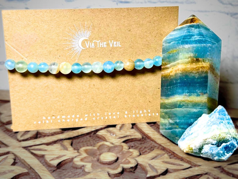 Puede incluir: Un brazalete de cristal azul claro con una luna creciente y el texto "Via The Veil" en una tarjeta marr&oacute;n. El brazalete est&aacute; hecho con cuentas azules claras y amarillas. La tarjeta tambi&eacute;n dice "Handmade with love & light, Reiki charged crystal bracelets".