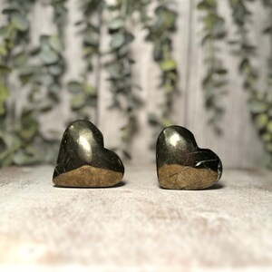 May include: Two gold pyrite heart shaped stones on a white wooden background.