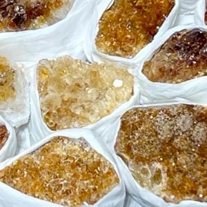 Op de afbeelding: Een verzameling ruwe citrienkristalclusters, elk afzonderlijk verpakt in wit papier. De kristallen vertonen verschillende tinten goudgeel, amber en dieporanje, met grillige, kristallijne oppervlakken en doorschijnende randen.