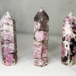 Pu&ograve; includere: Tre torri di cristallo lucido con punte. I cristalli hanno un mix di colori rosa, bianco e marrone scuro. I cristalli sono in piedi su una superficie bianca.