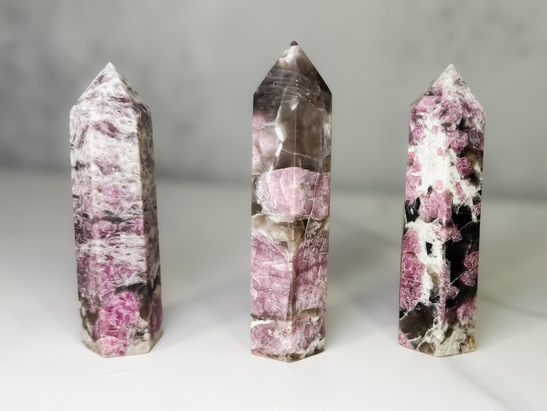 Pu&ograve; includere: Tre torri di cristallo lucido con punte. I cristalli hanno un mix di colori rosa, bianco e marrone scuro. I cristalli sono in piedi su una superficie bianca.