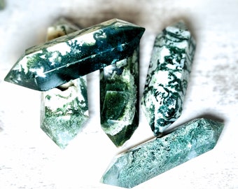 May include: Four green and white moss agate points, also known as dendrite agate, are arranged on a white wooden surface. The points are polished and have a natural, organic look.