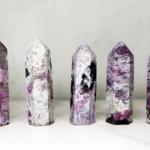 Pu&ograve; includere: Cinque torri di cristallo lucidate a forma esagonale. I cristalli mostrano un mix di tonalit&agrave; bianche, grigie e rosa. I cristalli sono in piedi su una superficie bianca, con uno sfondo grigio chiaro.