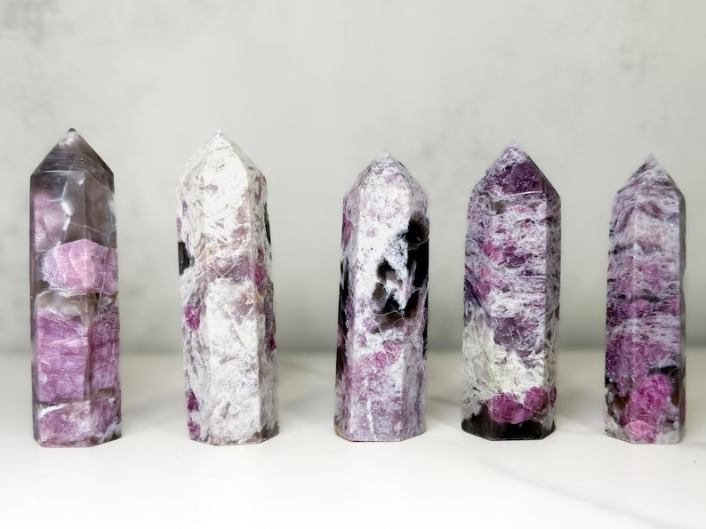 Pu&ograve; includere: Cinque torri di cristallo lucidate a forma esagonale. I cristalli mostrano un mix di tonalit&agrave; bianche, grigie e rosa. I cristalli sono in piedi su una superficie bianca, con uno sfondo grigio chiaro.