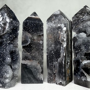 May include: Four polished crystal towers with pointed tops, featuring dark black and charcoal gray stone with rough, sparkling druzy crystal clusters embedded within the natural formations, standing upright in a row against a light background.