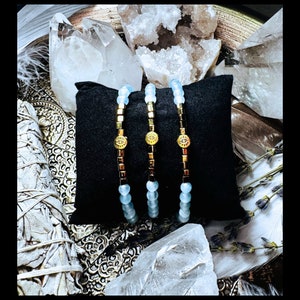 Puede incluir: Tres pulseras de cuentas doradas y azules con un dije de ojo turco. Las pulseras est&aacute;n sobre un coj&iacute;n de terciopelo negro y rodeadas de cristales blancos.