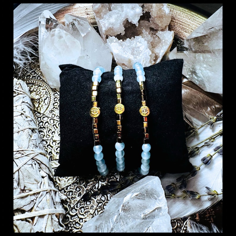 Puede incluir: Tres pulseras de cuentas doradas y azules con un dije de ojo turco. Las pulseras est&aacute;n sobre un coj&iacute;n de terciopelo negro y rodeadas de cristales blancos.