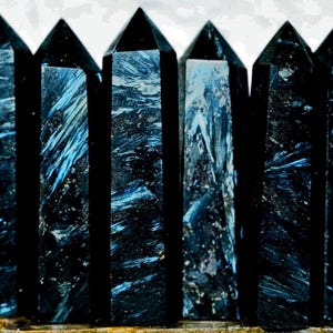 May include: Seven black and blue crystal points, also known as  "Blue Tiger's Eye" or "Hawk's Eye", are arranged in a row. The crystals are polished and have a smooth, glassy finish.
