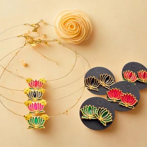 May include: Gold-toned lotus flower necklaces and earrings in various colors, including pink, black, red, and green. The earrings are displayed on black circular cards. A cream-colored rose is also present.