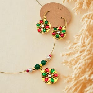 May include: A gold-toned jewelry set featuring a necklace and matching earrings. The earrings and pendant have a floral design with red, green, and white gemstones. The necklace has green beads and white pearls. The jewelry is displayed on a neutral background.