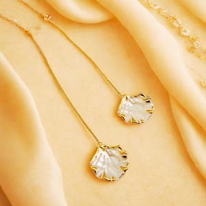 May include: A pair of delicate gold-toned drop earrings. Each earring features a white, petal-shaped pendant with gold trim. The earrings hang from fine gold chains, set against a soft, yellow background.