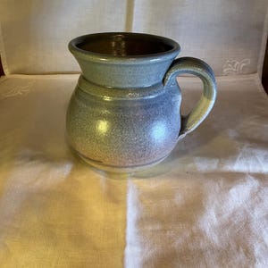 May include: A round ceramic mug with a wide, flared rim and a sturdy handle. The mug features a variegated glaze with soft blue, grey, and earthy brown tones, transitioning from a lighter blue at the top to a darker, speckled brown base.