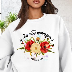 May include: White sweatshirt with a floral design and the text "do not worry" in black. The floral design includes red poppies and yellow flowers. The text "Matthew 6:34" is also included in black.