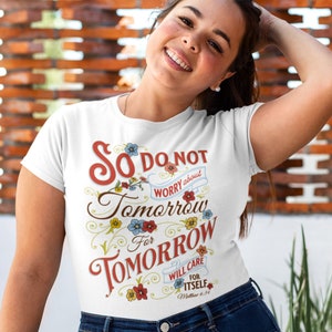 May include: White t-shirt with the text "So do not worry about tomorrow, for tomorrow will care for itself." The text is in red, brown, and blue, with floral accents. The shirt is a crew neck.