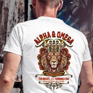 May include: White t-shirt with a graphic design featuring a lion wearing a crown and surrounded by roses. The text "ALPHA & OMEGA" is above the lion, and the text "LION OF THE TRIBE OF JUDAH THE BRIGHT AND MORNING STAR HE WHO WAS, WHO IS, AND IS TO COME" is below the lion.