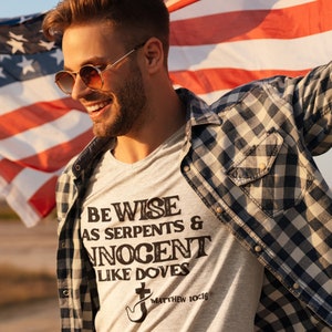 May include: A light gray t-shirt with the text "BE WISE AS SERPENTS & INNOCENT LIKE DOVES" and a dove graphic. The shirt is worn with a plaid shirt and sunglasses, and an American flag is in the background.