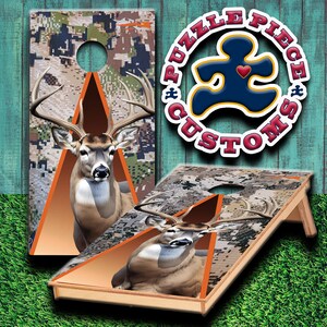 Könnte beinhalten: Zwei hölzerne Cornhole-Bretter mit einem Tarnmuster und einem Hirschbild. Die Bretter haben einen orangefarbenen Rand und ein dreieckiges Loch. Die Bretter befinden sich auf einer grünen Grasfläche. Die Bretter sind mit einem Logo verziert, auf dem "Puzzle Piece Customs" steht.