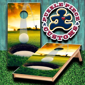 Puede incluir: Un juego de dos tablas de cornhole de madera con temática de golf. Las tablas están pintadas con una escena de puesta de sol y una bola de golf que entra en un agujero. Las tablas están decoradas con el texto "Puzzle Piece Customs".