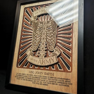 May include: Framed wooden artwork featuring a layered design of a rib cage and the text "THE BACK OF THE ARMY." The artwork includes the name "MSG JOHN SMITH" and a message of gratitude. The frame is black.