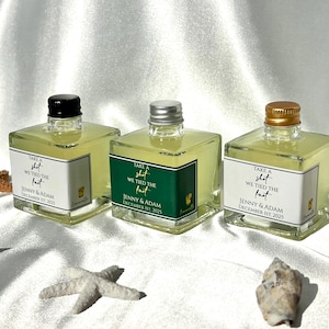 May include: Three square glass bottles filled with a yellow liquid, each with a different colored cap: black, silver, and gold. The bottles have labels that read "Take a shot, We tied the knot, Jenny & Adam, December 1st, 2025."