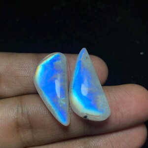以下が含まれることがあります： 2つの磨かれた、涙の形をしたムーンストーンの宝石。石は、青、紫、黄色の虹色の閃光を持つ乳白色の白いベースを示しています。宝石は暗い背景に対して手で保持されています。