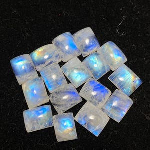 May include: A cluster of square-cut moonstone gemstones. Each stone has a milky white base with iridescent blue and rainbow flashes. The gemstones are displayed on a black surface, highlighting their natural beauty and suitability for jewellery.