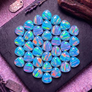 Pode incluir: Uma coleção de opalas cabochão de formato triangular dispostas em um grupo. Cada opala exibe uma cor de base azul-claro translúcida com flashes vibrantes de iridescência laranja, verde e amarela. As pedras são lisas e polidas.