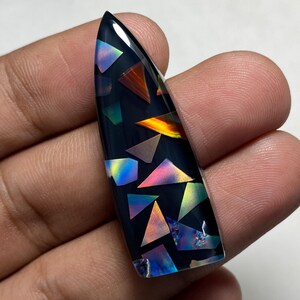 May include: A teardrop-shaped cabochon with a smooth, glossy black base embedded with various iridescent, multi-colored geometric shards. The surface features reflective triangular and irregular shapes that display a spectrum of colors including blue, orange, and pink hues.