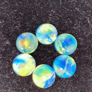 May include: Six iridescent glass cabochons arranged in a circle. The cabochons have a round shape and display a mix of blue, yellow, and green colors. The glass has a glossy finish, and the background is a dark, textured surface.