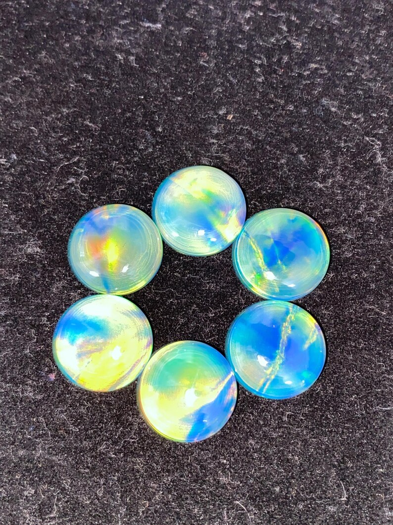 May include: Six iridescent glass cabochons arranged in a circle. The cabochons have a round shape and display a mix of blue, yellow, and green colors. The glass has a glossy finish, and the background is a dark, textured surface.