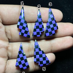 Op de afbeelding: Vijf driehoekige edelsteenhangers met een zwart-paars dambordpatroon. Elke hanger heeft een puntige top en een unieke vorm. De edelstenen worden op een hand tegen een donkere achtergrond getoond.