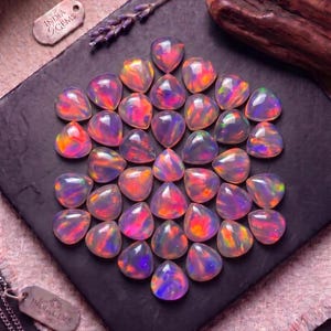 Puede incluir: Una colección de ópalos en forma de pera dispuestos en un patrón hexagonal sobre una superficie de pizarra negra. Cada gema muestra un cuerpo translúcido con destellos vibrantes de fuego rosa, naranja y verde. Dos etiquetas metálicas INDIA GEMS son visibles.