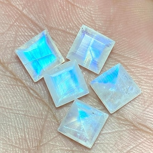 May include: Five square-cut moonstone gemstones with iridescent blue and white flashes. The gemstones are displayed on a light-colored surface, showcasing their polished facets and natural beauty. These stones are ideal for jewelry making.