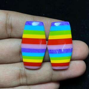 Könnte beinhalten: Zwei fassförmige Perlen mit einem Regenbogenstreifen-Design. Die Perlen zeigen ein Farbspektrum, darunter Lila, Blau, Grün, Gelb und Rot. Die Enden der Perlen sind hellrosa.