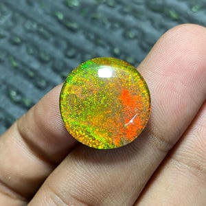 May include: A round, polished gemstone with a vibrant display of color. The stone features a mix of green, orange, and yellow hues, creating a fiery and iridescent effect. The gemstone is held in a hand, showcasing its size and shape.